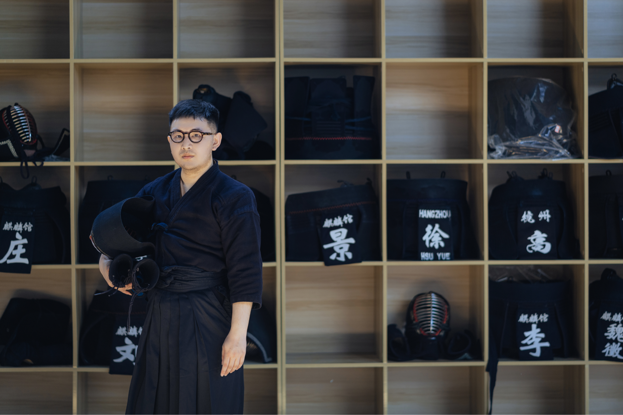 Kyle in kendo uniform, holding a helmet, in front of a wall of labelled equipment shelves